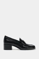 Mocasín de mujer en color negro con acabado liso, diseño de punta cuadrada y tacón en bloque. Presenta un detalle metálico decorativo en el antifaz frontal.
