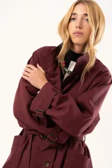 Trench de corte holgado color bordó, con cuello camisero, cierre frontal con botones y cinturón ajustable con hebilla. Presenta mangas largas con detalles de correas en los puños y bolsillos laterales.