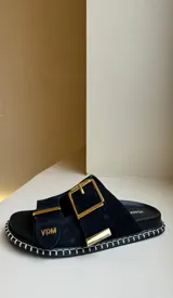 Sandalias planas estilo birkenstock de nobuck negro, con dos tiras ajustables y hebillas metálicas doradas. La suela tiene un pespunte decorativo blanco alrededor del borde.