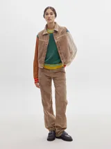 Chaleco de pana negro con cierre frontal de botones metálicos dorados. Presenta detalles de costuras en contraste en el cuello, cartera y canesú, e incluye un pequeño parche con forma de bandera en el pecho.