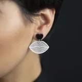 Par de aros colgantes con forma de labios delineados en blanco y rellenos de glitter plateado, con un corazón negro en la parte superior.