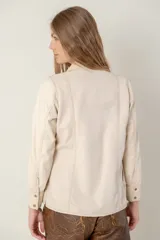 Camisa de cuero ovino color beige claro, estilo western, con canesú en los hombros en gamuza de tono similar. Presenta costuras contrastantes y cierre frontal con broches. Posee bolsillos laterales.
