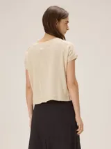 Remera tejida color beige de corte relajado, con mangas cortas y detalle de costuras frontales marcadas. Incluye un pequeño bordado distintivo en el borde inferior.