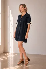 Vestido corto azul marino de lino, con cuello polo, cierre frontal parcial de botones y mangas cortas con detalles de lazos en color tostado. Presenta costuras a contratono y bolsillos laterales.