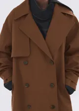 Chaqueta larga tipo trench color marrón, con diseño recto, solapas anchas, doble botonadura, bolsillos laterales y cinturón desmontable de la misma tela.