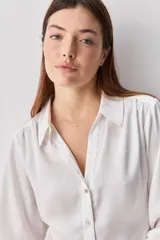 Camisa blanca de manga larga con textura suave y caída liviana, cuello camisero, puños anchos y fruncidos sutiles en los hombros.