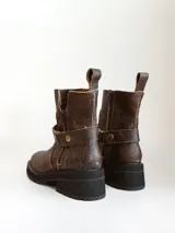 Bota corta de cuero vacuno en color chocolate con efecto desgastado. Presenta una suela track de goma negra, detalle de pulsera desmontable con tachas metálicas y cierre lateral.