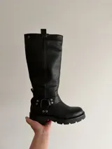 Bota de caña alta confeccionada en cuero sintético, con suela track robusta y detalle de arnés con argolla metálica y remaches en la zona del tobillo.