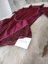 Body de lycra color bordó con cuello alto, mangas cortas y espalda descubierta. Presenta detalles de encaje en el cuello, mangas y laterales de la parte inferior. Cuenta con cierre de broches en la entrepierna.