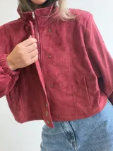 Chaqueta corta de efecto gamuza en color azul marino, con cierre frontal y solapa decorada con una hilera de aros metálicos. Presenta mangas tipo globo, ajuste lateral en la cintura y forro interior.