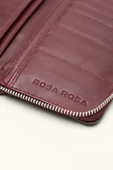 Billetera rectangular de cuero con textura de reptil, cierre perimetral y correa de muñeca lateral. Presenta un pequeño aplique metálico circular en el centro.