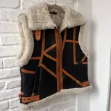 Chaleco negro de cuero con interior de lana color crema, cierre frontal con cremallera y detalles de tiras de cuero marrón.