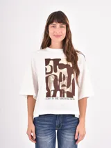 Remera de corte oversize color blanco con cuello redondo y mangas cortas. Presenta un estampado frontal rectangular con formas abstractas en tonos marrón y dorado, acompañado por la frase 'LOST IN THE ABSTRACTION'.