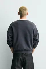 Sudadera de corte boxy fit en color gris oscuro con efecto lavado. Presenta cuello redondo en contraste color crema, manga larga con puños acanalados y estampado gráfico frontal con estrellas y tipografía estilo vintage.
