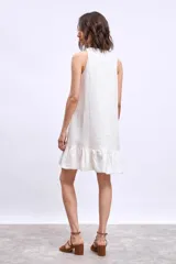 Vestido corto blanco sin mangas, con escote en V y detalle de broche dorado en el centro. La falda tiene un volado en el ruedo.