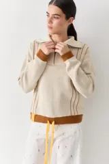 Buzo de punto color beige con rayas verticales marrones, cuello polo con cierre frontal, puños y ruedo acanalados en color marrón. Incluye cordones amarillos ajustables en la cintura.