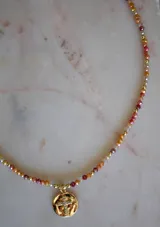 Collar de cristales facetados en tonos naranja, rojo y beige, con esferas doradas intercaladas. Incluye un dije redondo de baño de oro con una cruz de circonitas incrustadas.