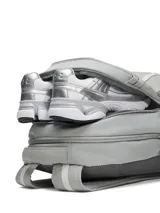 Mochila Under Armour modelo Hustle 6.0, color gris, con tecnología UA Storm resistente al agua. Cuenta con compartimento acolchado para portátil, bolsillo para calzado o ropa sucia, bolsillos laterales de malla y sistema Molle para sujeción.
