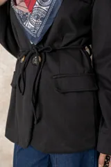 Chaquetón color beige de corte recto y largo, con cierre frontal mediante botones y lazo ajustable en la cintura. Presenta cuello en V, bolsillos laterales con solapa y mangas largas.