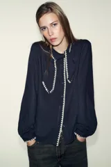 Blusa de manga larga en color azul marino, con cuello solapa y lazo frontal. Presenta detalles de pespuntes bordados en color crema a lo largo del cuello, lazo, cierre frontal y puños. Cierre frontal con botones.