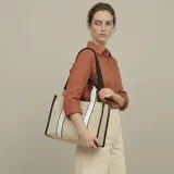 Bolso tipo tote bag de lona color beige con detalles en negro y dos franjas verticales blancas en el frente. Cuenta con asas de mano y una correa ajustable para llevar al hombro.