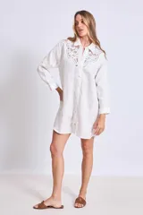 Vestido camisero corto color crudo, de lino, con cuello camisero, mangas largas y cierre frontal con botones. Presenta apliques de encaje en cuello y hombros.