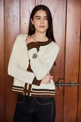 Cardigan de punto color crudo con detalles en marrón y negro. Presenta cuello redondo, cierre frontal con botones forrados y puños acanalados con rayas horizontales.