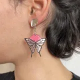 Par de aros colgantes con forma de mariposa plateada con detalles en negro y círculo superior rosa.