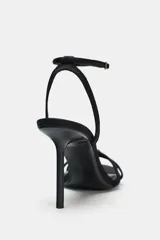 Sandalia de tacón alto fino, color negro, con tiras finas cruzadas en el empeine y un detalle de abalorio rojo y dorado en la unión de las tiras. Posee tira ajustable con hebilla en el tobillo.