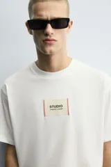 Camiseta boxy fit de color blanco con cuello redondo y mangas cortas. Presenta un parche rectangular en el centro del pecho con texto impreso y costuras decorativas a contraste en los laterales.