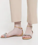 Sandalia plana color nude con tira fina con aplicación de strass y mini espejos, cierre en el tobillo, puntera redonda y taco bajo plano.