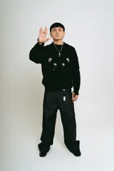 Pantalón de jean negro, corte baggy, confeccionado en denim crudo. Presenta un estampado serigrafiado en blanco con un diseño estilizado en ambos bolsillos traseros y un parche con logo en la cintura.