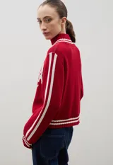 Campera tejida de punto color marrón con cierre frontal completo, cuello alto y detalles de rayas blancas en cuello, puños y ruedo. Presenta el logo de la marca estampado en el frente.