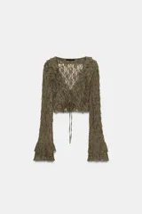 Chaqueta corta de tejido tipo encaje color verde oliva, con escote pico, manga larga acampanada y detalle de volantes en el borde del escote y los puños. Cierre frontal mediante lazadas.