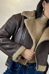 Campera tipo aviador de cuero sintético marrón con interior y detalles de corderito beige. Presenta cierre frontal cruzado, cuello solapa amplio, puños con corderito y cinturón ajustable en la cintura con hebilla metálica.