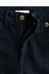 Jean tobillero de denim azul oscuro, de tiro alto y piernas amplias con corte holgado. Presenta bolsillos delanteros tipo parche con solapa y botones dorados, además de pliegues marcados en el frente.