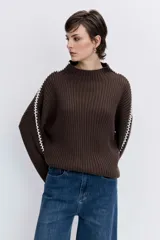 Jersey de punto color marrón chocolate, de cuello alto y corte holgado. Presenta un diseño acanalado y un detalle de costura decorativa en color blanco que recorre la parte superior de las mangas.