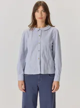 Camisa de denim con silueta levemente entallada, cuello bebé y mangas largas con sutil frunce en los hombros. Presenta costuras curvas decorativas con pespuntes en contraste y cierre frontal con botones.