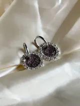 Aros colgantes con forma circular, hechos de resina color lila y borde de strass plateado. El aro es de acero quirúrgico plateado.