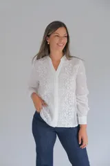 Blusa blanca de manga larga con cuello mao y cierre frontal de botones ocultos. Presenta un delicado diseño con bordados florales y detalles calados verticales en el frente.