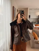 Chaqueta marrón de cuero con cuello de piel sintética negra, cierre con botones a presión, bolsillos con solapa y corte a la cintura.