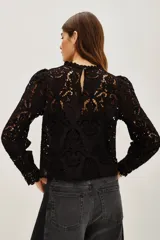 Camisa de encaje negro con cuello alto y mangas largas abullonadas. Presenta un diseño calado con motivos florales y cierre posterior.