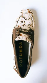 Mocasín tipo náutico de cuero con pelo, con diseño estampado de manchas en tonos marrones y blancos. Presenta detalles en gamuza marrón en el talón y la zona de los cordones, con ojalillos metálicos laterales.