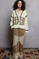 Cardigan de tejido de punto grueso color crema, con cuello tipo camisa y cierre frontal de botones florales. Presenta detalles deshilachados y teñido tipo tie-dye en tonos grises oscuros en el frente y puños. Tiene parches cuadrados de tela a cuadros en tonos tierra sobre los bolsillos delanteros.