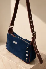 Cartera tipo bandolera de gamuza marrón, con detalles de tachas y ojalillos metálicos en el frente. Cuenta con cierre superior, bolsillo trasero con cierre, correa ajustable y placa metálica con la marca grabada.