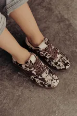 Zapatillas de cuero con estampado que simula pelo de vaca o manchas marrones y blancas. Tienen detalles en cuero marrón liso, cordones marrones y suela de goma color caramelo.