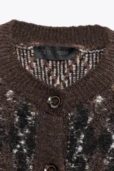 Chaqueta de punto texturizado con cuello redondo y manga corta. Presenta un patrón de rombos en tonos marrón oscuro, negro y blanco. Cierre frontal con botones oscuros.