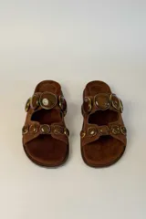 Sandalias planas de gamuza marrón, con dos tiras anchas adornadas con grandes piedras redondas facetadas en tonos marrones con bordes metálicos dorados.
