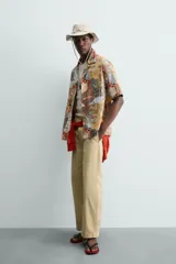 Camisa de corte relajado confeccionada en tejido ligero de algodón, con cuello bowling y manga corta. Presenta un estampado integral con motivos urbanos y tropicales en tonos cálidos.