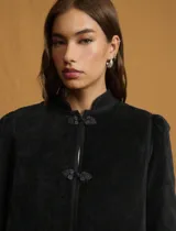 Chaqueta negra de estilo oriental con cuello alto estructurado y cierre frontal mediante nudos decorativos tradicionales. Presenta mangas largas con leve volumen en los hombros y un corte recto.